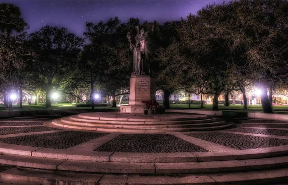Moody night scene of a bronze statue on a circular stone plaza with tiered steps, oak trees and glowing lampposts under a purple sky in a quiet city park.