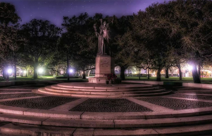Moody night scene of a bronze statue on a circular stone plaza with tiered steps, oak trees and glowing lampposts under a purple sky in a quiet city park.
