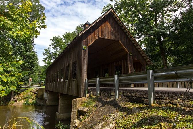 Quaint rustic wooden covered bridge spanning a creek on stone supports, flanked by trees and a metal guardrail in a sunlit rural setting