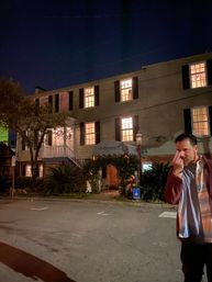Nighttime scene of a historic downtown colonial-style building with warm lit windows and a decorated entrance arch; a casually dressed man stands in the foreground making a playful hand gesture.