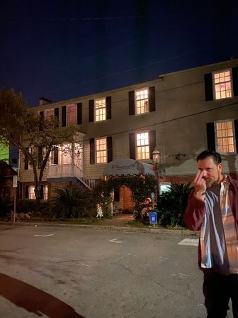 Nighttime scene of a historic downtown colonial-style building with warm lit windows and a decorated entrance arch; a casually dressed man stands in the foreground making a playful hand gesture.