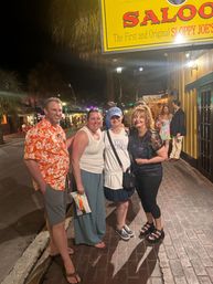 Four friends smiling on a brick sidewalk at night beneath a yellow saloon sign, palm trees and colorful lit storefronts in a lively downtown nightlife scene
