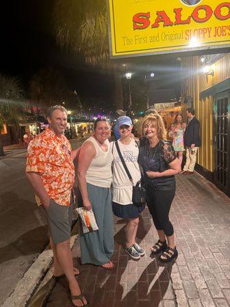Four friends smiling on a brick sidewalk at night beneath a yellow saloon sign, palm trees and colorful lit storefronts in a lively downtown nightlife scene