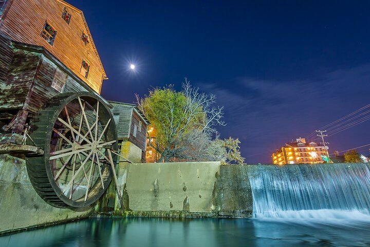 Historic wooden watermill with a giant water wheel beside a concrete spillway and silky waterfall under a bright moon, lit trees and distant glowing buildings reflecting in the calm river