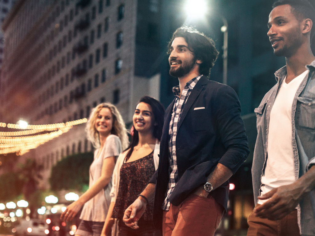 Four friends smiling as they walk down a lively downtown street at night, urban nightlife with glowing string lights and building facades in the background.