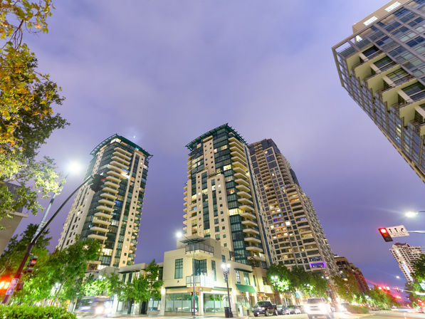Upward view of modern downtown high-rise apartment towers glowing at dusk, with lit balconies, street-level shops, trees and cars on a busy city street under a purple evening sky.