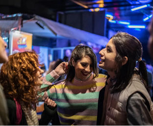 Three friends laughing and dancing at a lively outdoor city night market under colorful neon lights