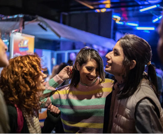 Three friends laughing and dancing at a lively outdoor city night market under colorful neon lights