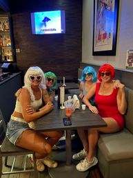 Four friends in colorful wigs and sunglasses posing at a casual bar booth with drinks on the table and a TV on the wall — playful night out.