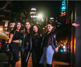 Four friends smiling on a downtown city street at night, neon signs and city lights behind them, enjoying a night out.