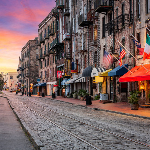 Sunset-lit historic cobblestone street in a Southern coastal city, lined with brick warehouse buildings, colorful awnings and flags, outdoor planters and vintage streetcar tracks leading into the distance.