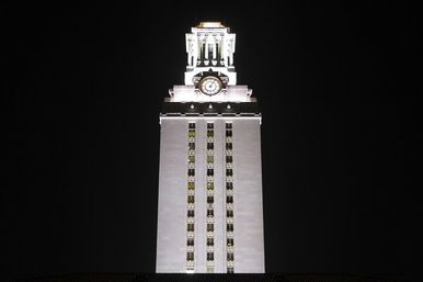 Illuminated white clock tower at night with a tall rectangular facade, vertical rows of windows, ornate lit belfry and prominent clock face against a black sky