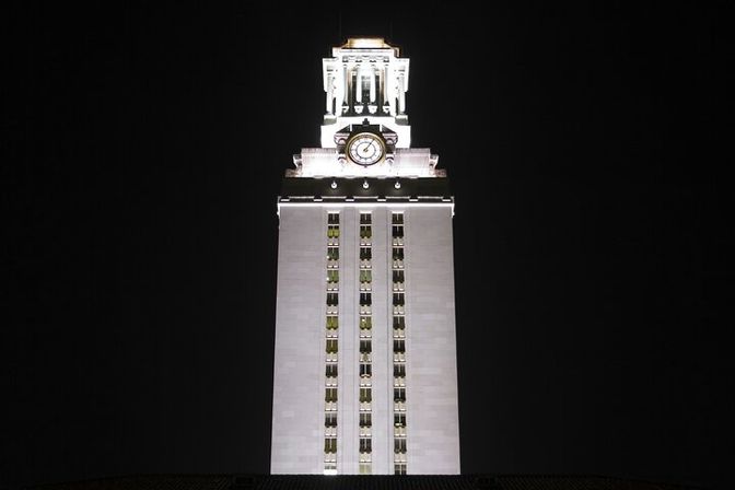 Illuminated white clock tower at night with a tall rectangular facade, vertical rows of windows, ornate lit belfry and prominent clock face against a black sky