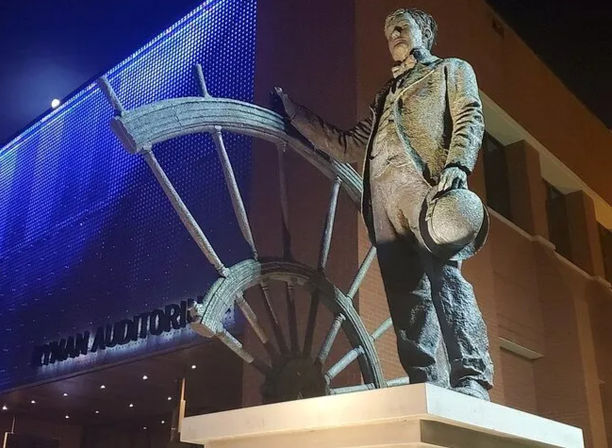 Bronze statue of a 19th-century–dressed man holding a hat and gesturing beside a giant wheel sculpture at the illuminated auditorium entrance at night.