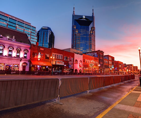 Sunset view of Nashville's Broadway entertainment district with neon-lit honky-tonk bars along the street and a dramatic glass skyscraper towering in the background.
