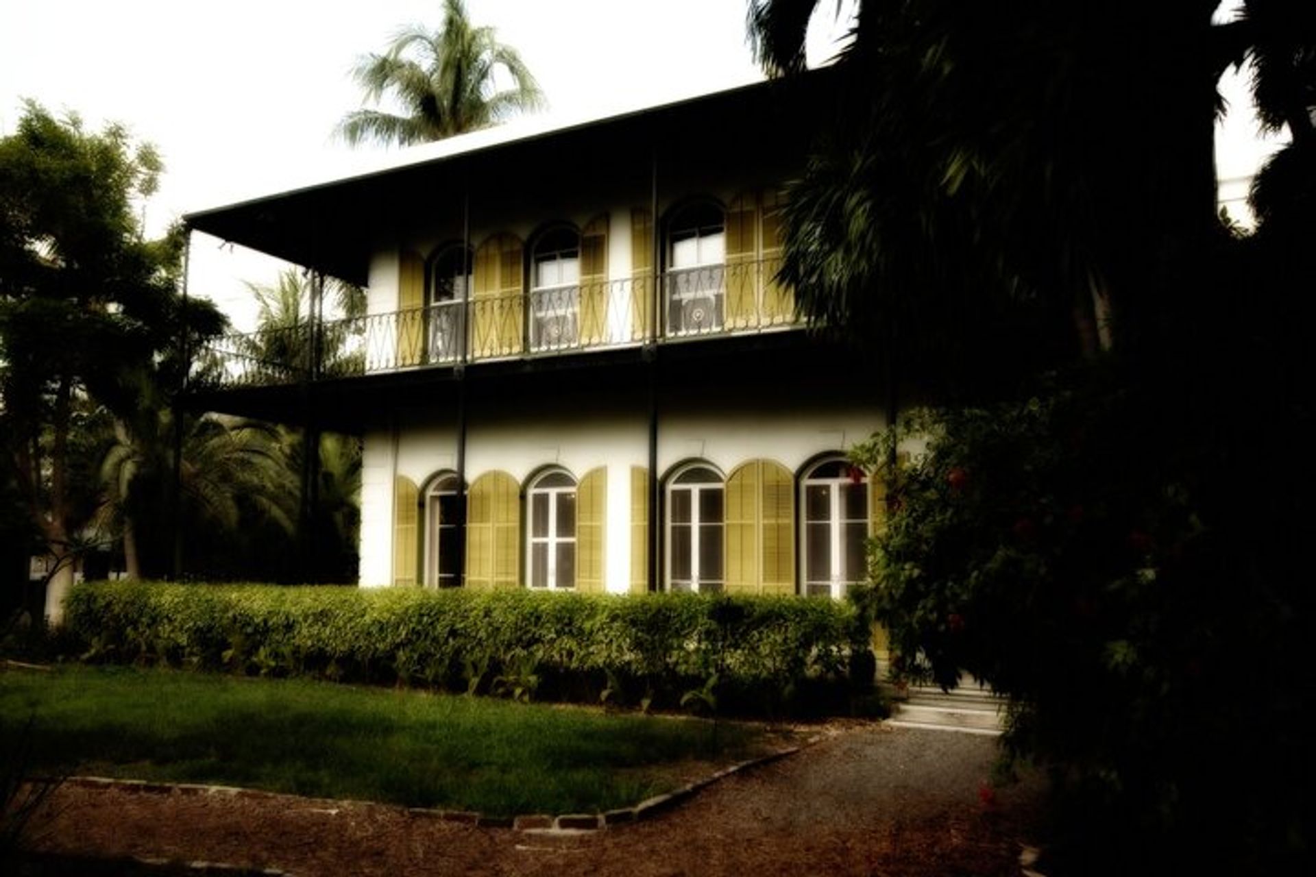 Charming two-story colonial-style house with arched shuttered windows and an iron balcony, framed by palm trees, hedges, and a gravel path in a tropical garden.