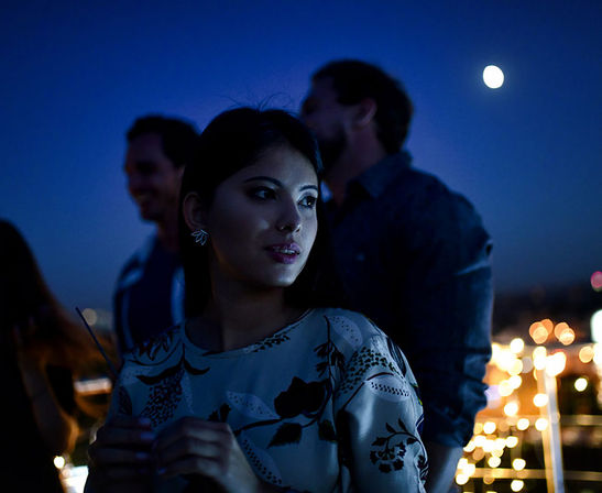 Young woman in a floral top at a moonlit rooftop party, gazing toward blurred guests and sparkling city lights in the night sky.