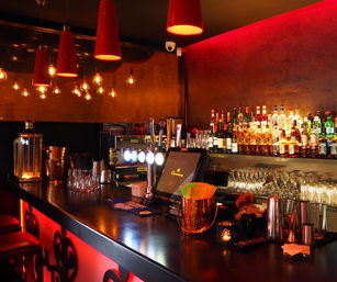 Moody red-lit cocktail bar interior with glossy counter, hanging pendant lamps and Edison bulbs, beer taps, rows of liquor bottles and glassware, bar tools and colorful straws—cozy nightlife vibe.