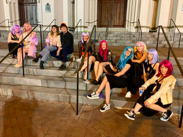 Around ten friends in colorful wigs and party outfits sitting and laughing on stone steps outside a city building entrance at night, posing for a group photo.