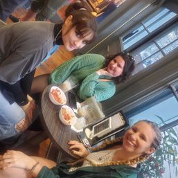 Three friends smiling at a small round table in a cozy downtown cafe by large windows, bowls with snacks and iced drinks on the table and city street visible outside.
