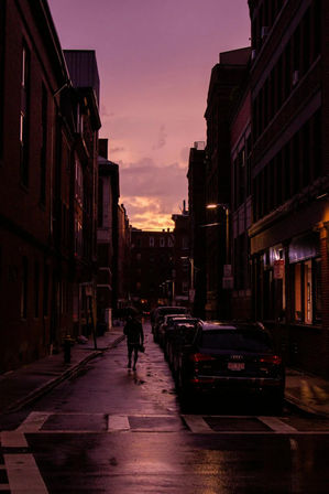 Rain-slicked downtown street at purple sunset, narrow brick-lined city alley with parked cars, glowing streetlights and a silhouetted pedestrian holding an umbrella.