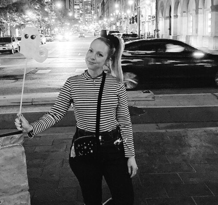 Person in a striped shirt holding a ghost-shaped balloon on a downtown city sidewalk at night, with a blurred car and illuminated storefronts in the background.