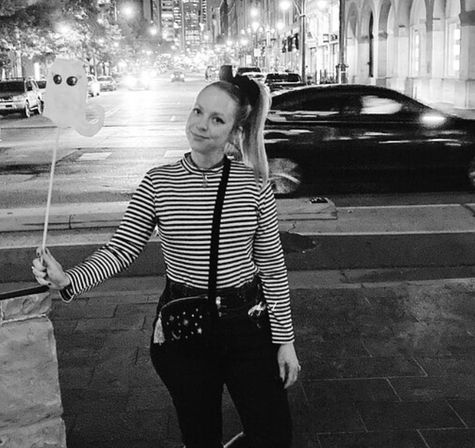 Person in a striped shirt holding a ghost-shaped balloon on a downtown city sidewalk at night, with a blurred car and illuminated storefronts in the background.