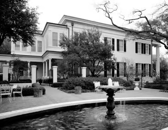 Black-and-white photo of an elegant two-story colonial-style mansion with shuttered windows, a wraparound porch, wrought-iron fence, courtyard fountain, outdoor seating, and large oak trees.