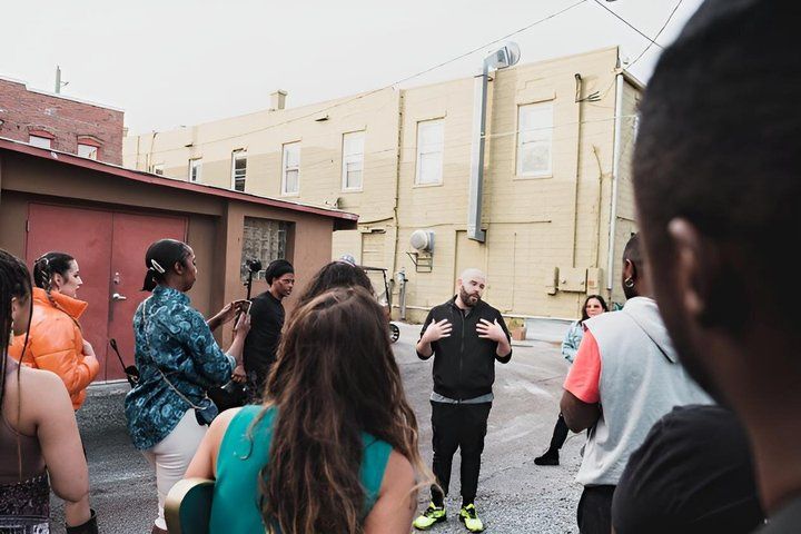 Urban alley scene: a casual speaker gestures to a small crowd of young adults—some filming on phones and one with a guitar—in front of yellow commercial buildings, street-side meetup vibes.