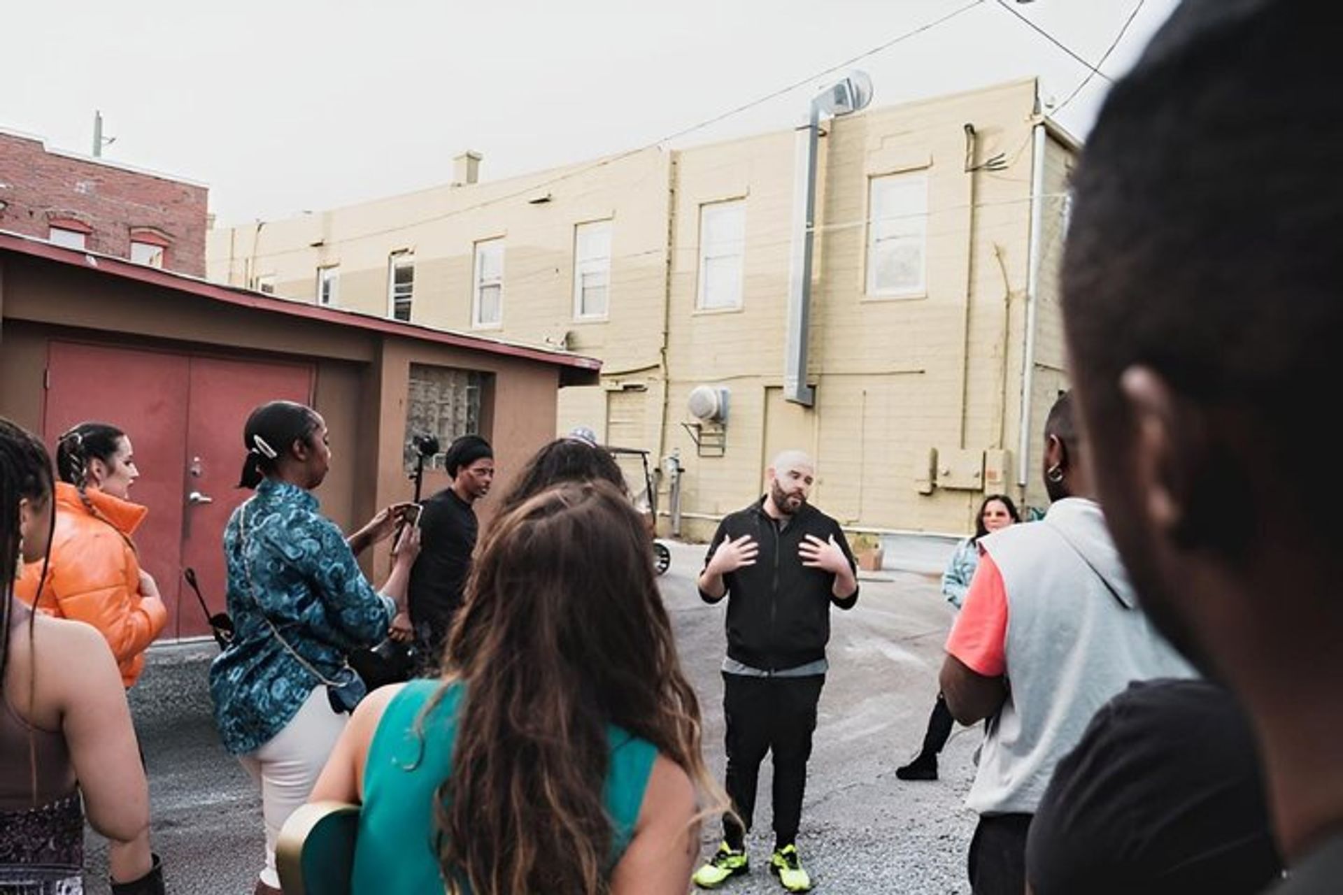 Urban alley scene: a casual speaker gestures to a small crowd of young adults—some filming on phones and one with a guitar—in front of yellow commercial buildings, street-side meetup vibes.