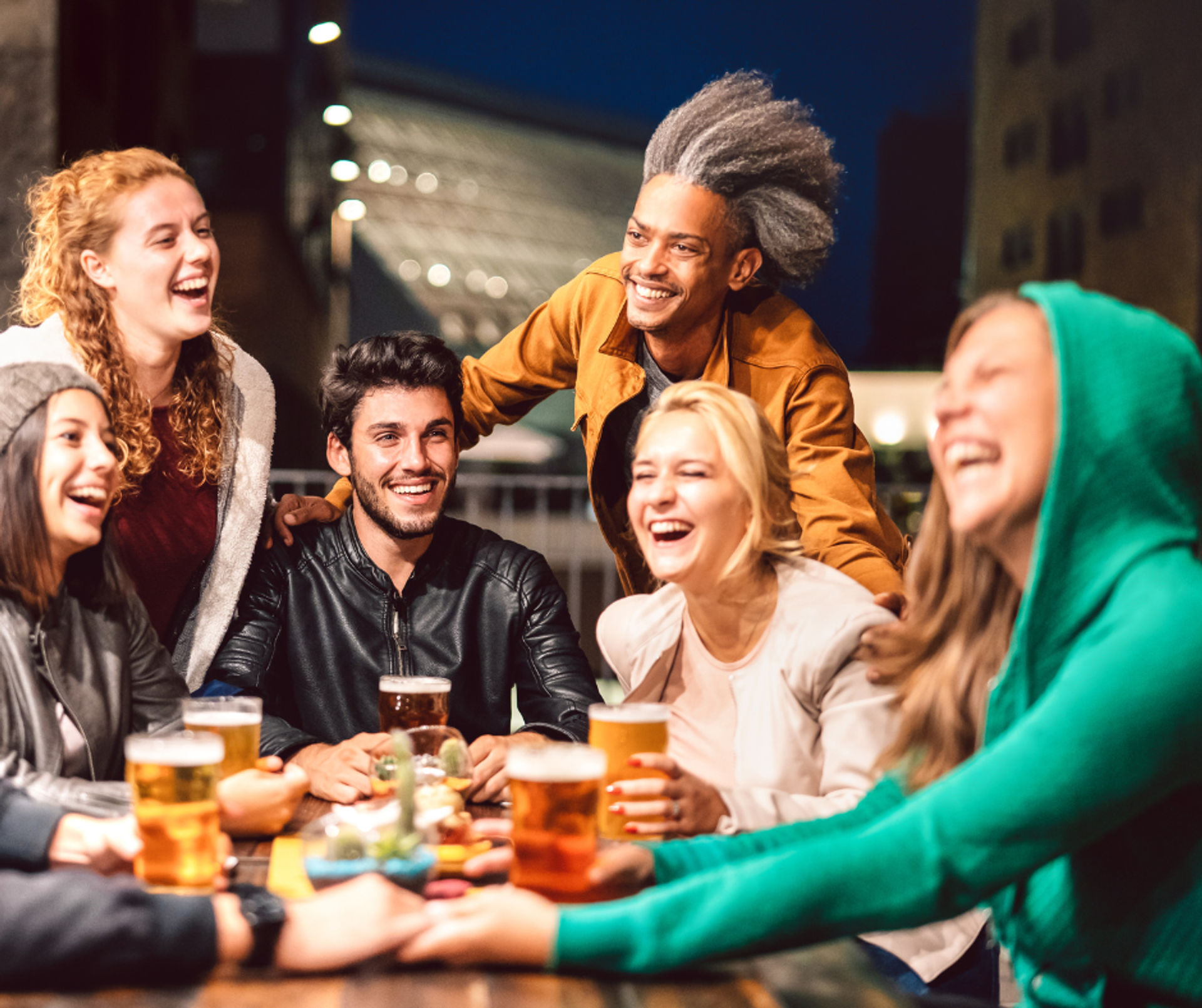 Six friends laughing and sharing pints around a wooden table on a city outdoor patio at night