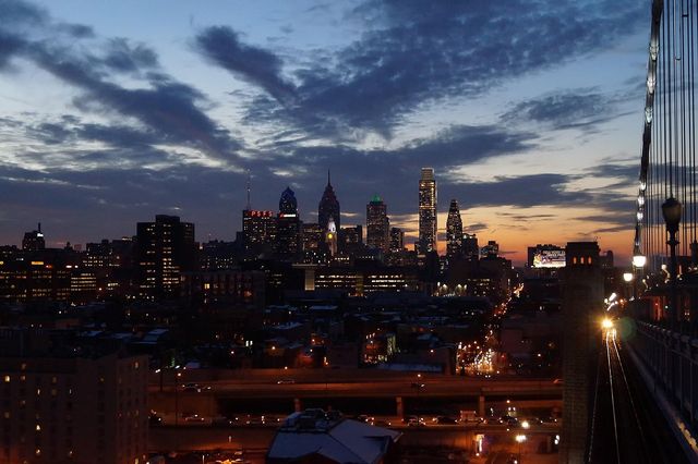 Philadelphia skyline at dusk seen from a bridge, glowing skyscrapers and city lights with streaks of traffic under a dramatic clouded sunset.
