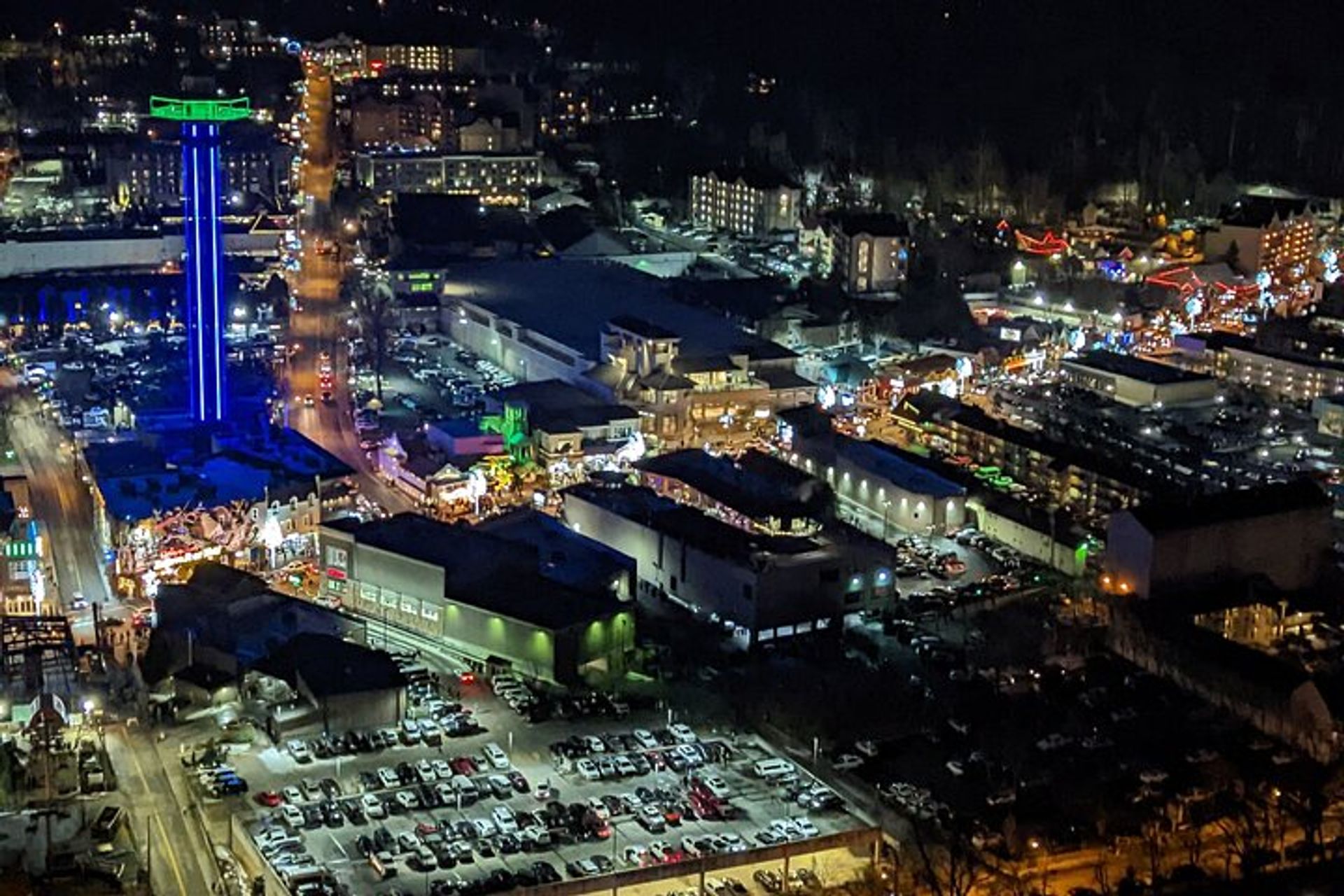 Aerial night view of a bustling mountain tourist district with a glowing blue observation tower, festive string lights, busy streets and a crowded parking lot.