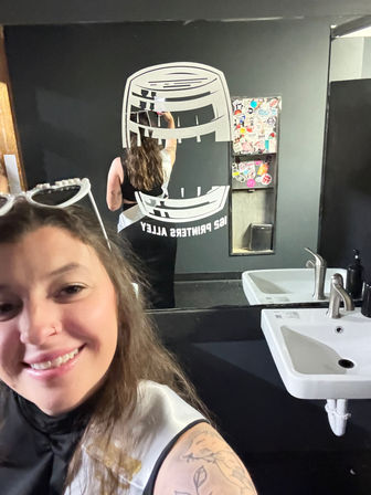 Smiling person taking a mirror selfie in a black-painted restroom, reflection shows a large white barrel-style wall decal, a sticker-covered dispenser, and a white sink with chrome faucet; tattooed arm visible.