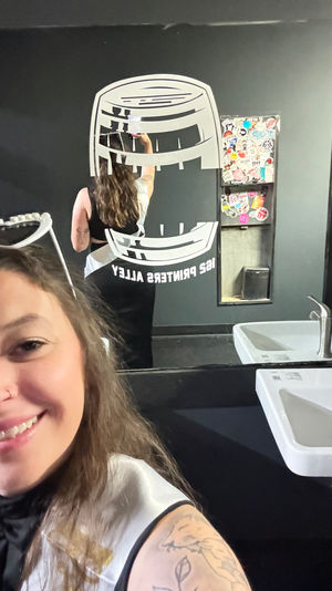Smiling person taking a mirror selfie in a black-painted restroom, reflection shows a large white barrel-style wall decal, a sticker-covered dispenser, and a white sink with chrome faucet; tattooed arm visible.