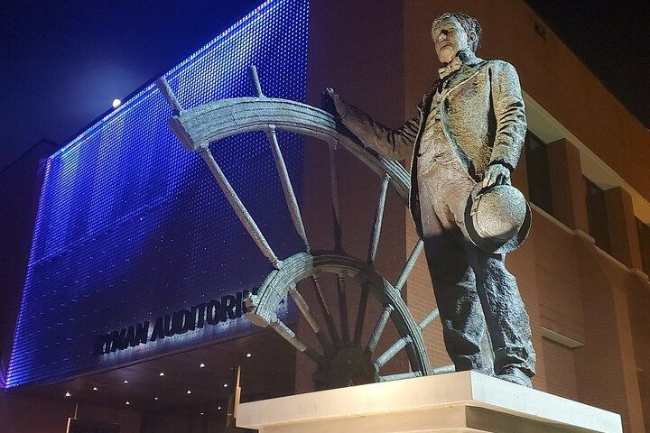 Bronze statue of a suited man holding a hat beside an oversized ship's wheel, lit by blue LED lights on an auditorium facade at night.