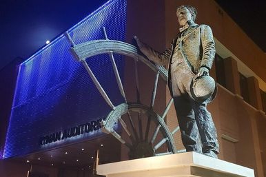 Bronze statue of a suited man holding a hat beside an oversized ship's wheel, lit by blue LED lights on an auditorium facade at night.