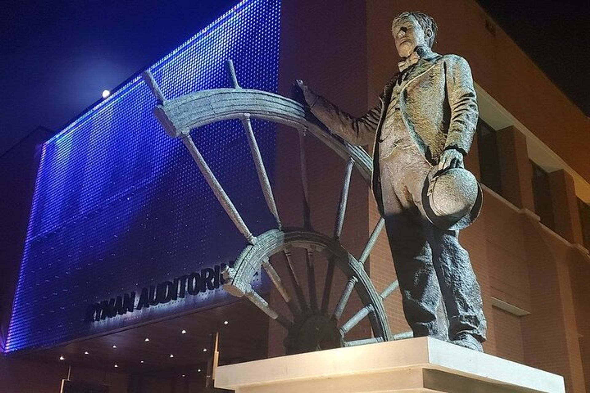 Bronze statue of a man in early 20th-century attire holding a hat beside a large wagon wheel on a pedestal outside a brick auditorium with blue-purple LED-lit facade at night.