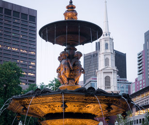 Ornate bronze tiered fountain with cherubic statues and streams of water, lit at dusk in a downtown square with a white church steeple and modern office buildings in the background.