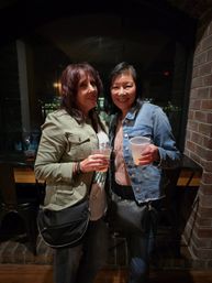 Two women smiling and holding drinks in plastic cups inside a cozy dim waterfront bar with exposed brick and a large window showing city lights at night.