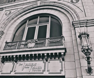 Black-and-white close-up of a historic stone building facade with a large arched window, ornate iron balcony and decorative crest, carved cornice details and a vintage wall lantern.