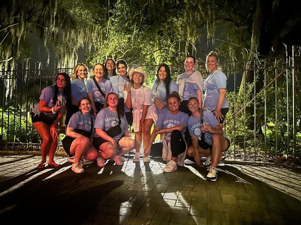 Smiling group of friends in matching light-blue 'Dancing Queens' shirts posing at night under moss-draped live oak in front of a wrought-iron fence.