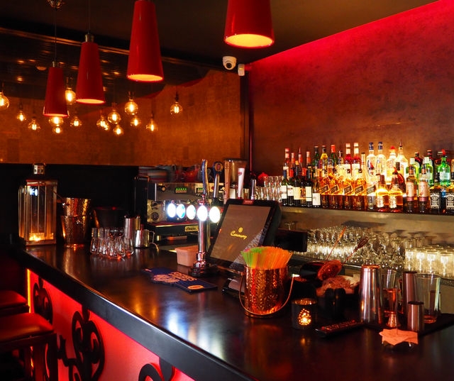 Dimly lit cocktail bar interior with red pendant lights, glowing liquor bottles on back shelves, glossy counter with draft taps, glassware and bar tools.