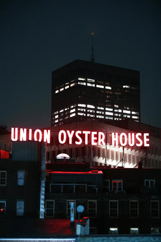 Night cityscape: glowing red neon rooftop sign for a seafood restaurant crowns brick rooftops, with a lit office tower looming against the dark sky.