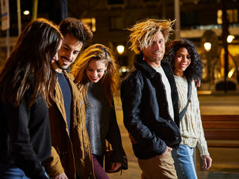 Five friends strolling and laughing down a city sidewalk at night under warm streetlights, wearing casual jackets and cozy sweaters.