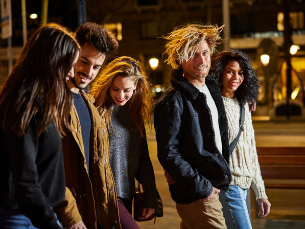 Five friends strolling and laughing down a city sidewalk at night under warm streetlights, wearing casual jackets and cozy sweaters.