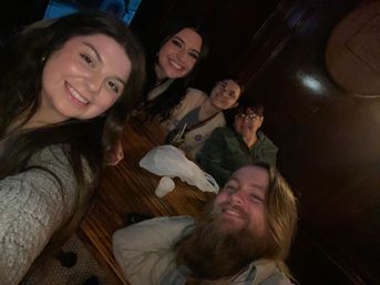 Five friends smiling for a selfie in a cozy, dim wood‑paneled pub around a wooden table with drinks — casual night out vibe.