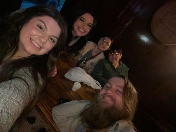 Five friends smiling for a selfie in a cozy, dim wood‑paneled pub around a wooden table with drinks — casual night out vibe.