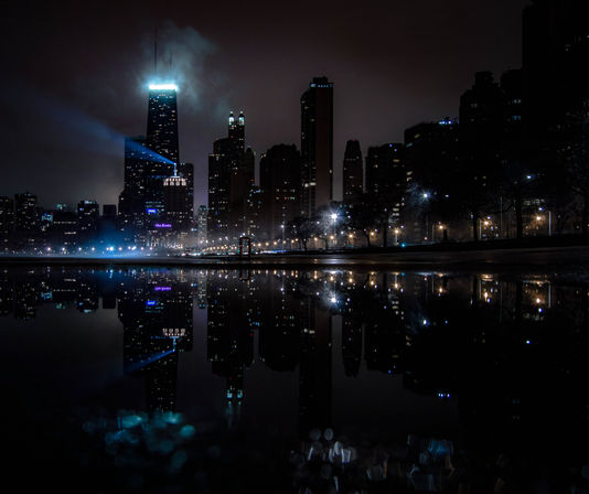 Moody waterfront city skyline at night with blue searchlight, glowing streetlights and sharp reflection in still water