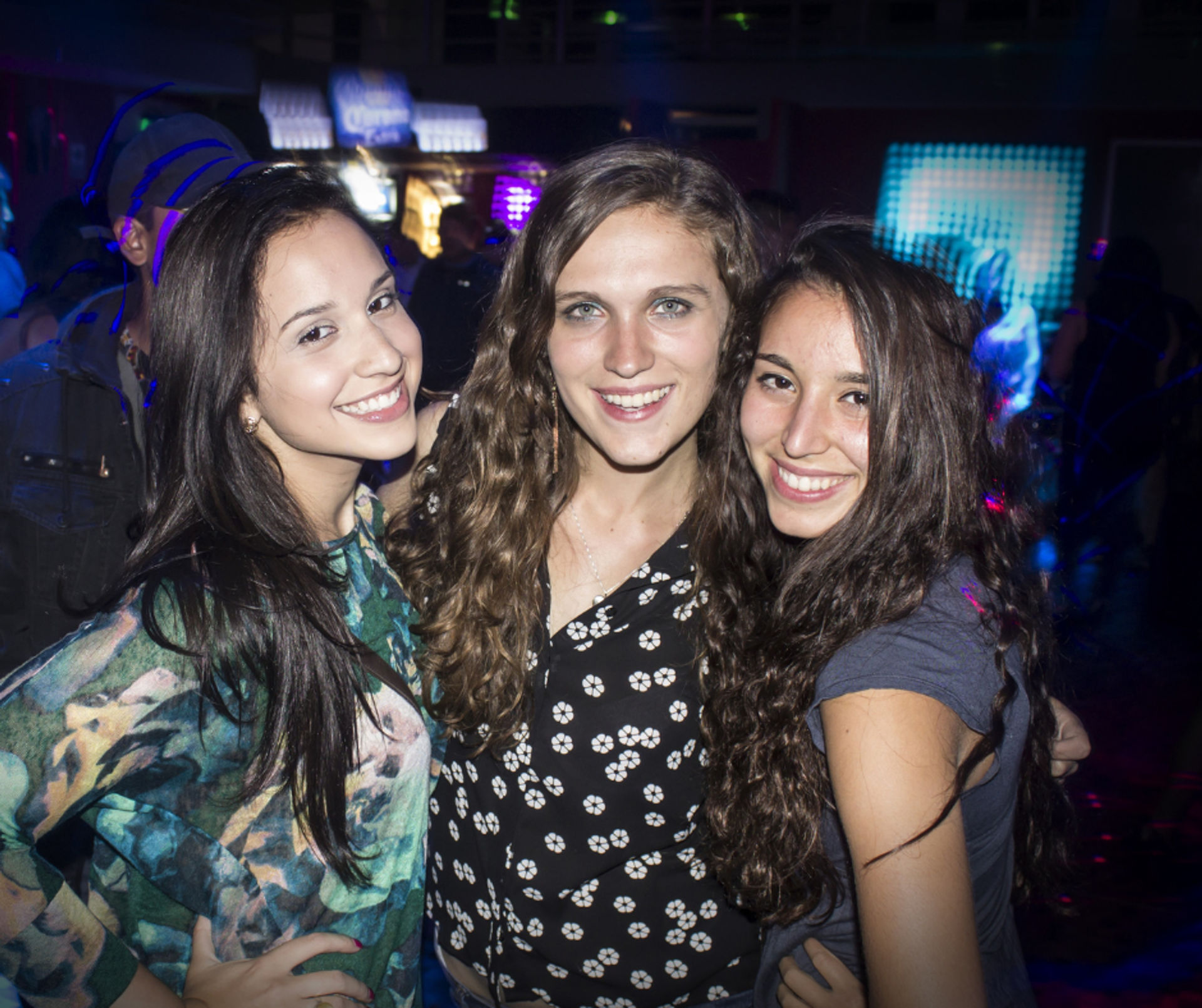 Three friends smiling and posing on a lively nightclub dance floor with colorful LED lights and a partying crowd in the background.